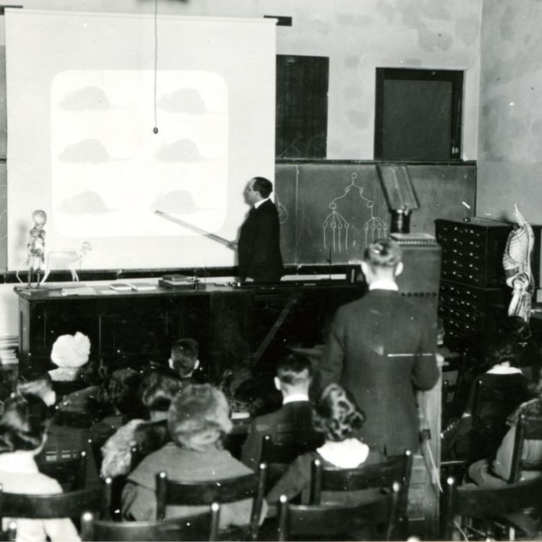 Black and white image. Teacher stands in the front of a classroom with a pointer.
